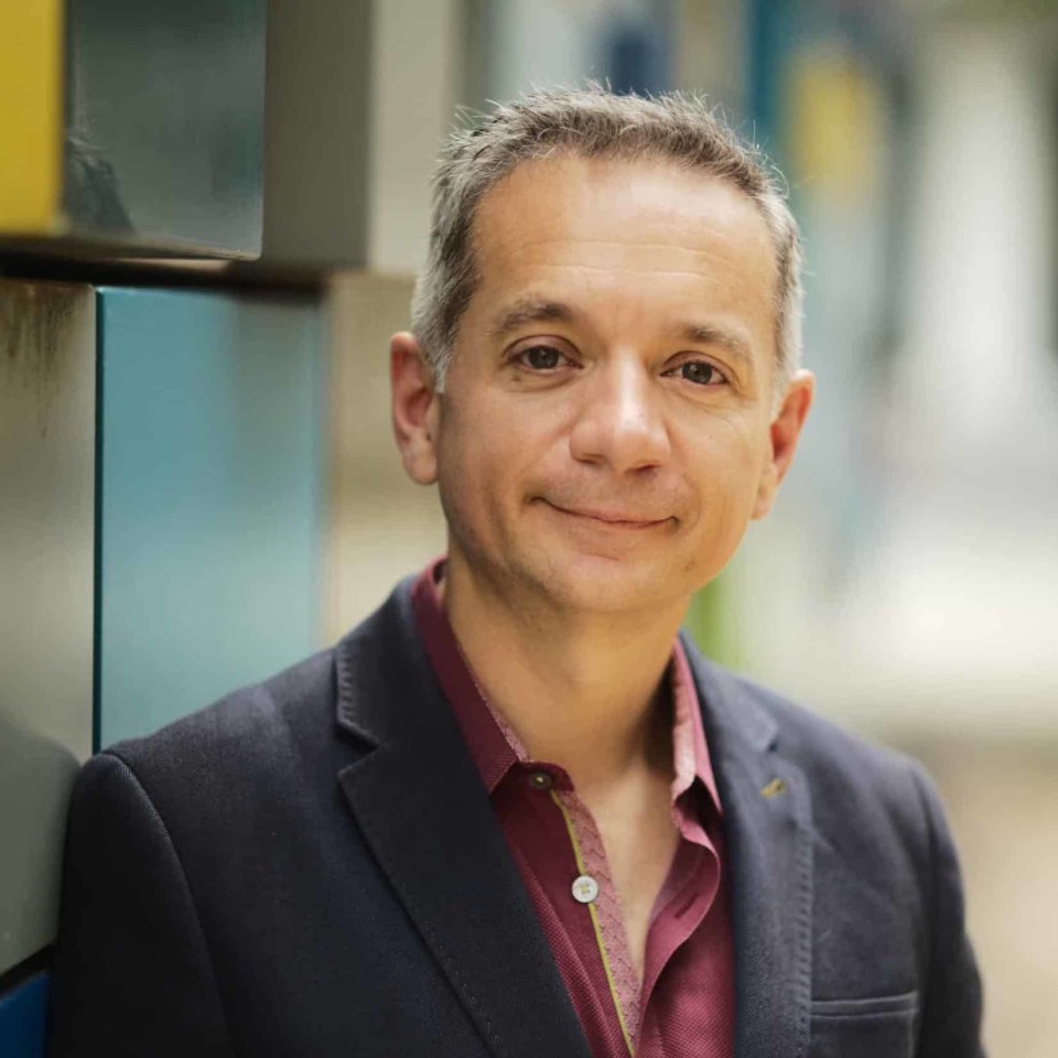 A smiling man with greying hair, wearing a dark blazer over a raspberry-colored shirt, posing for a portrait with a softly blurred background.