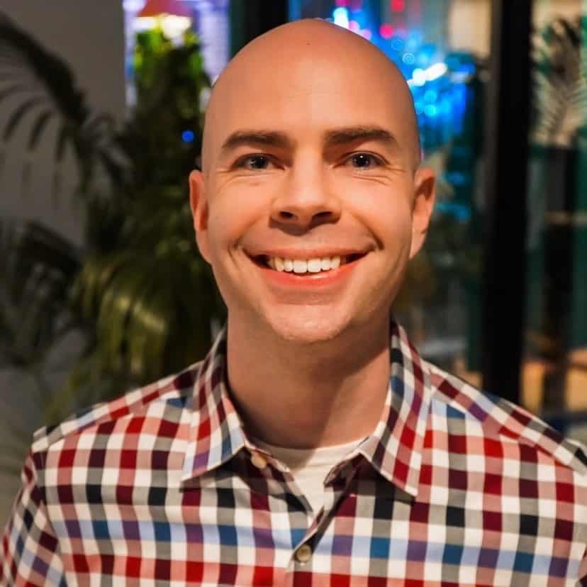 A smiling person with a shaved head, wearing a checkered shirt in a warmly-lit indoor setting.