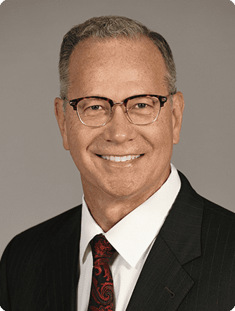 A professional headshot of a smiling middle-aged man wearing glasses, a dark suit, a red patterned tie, and a white shirt.
