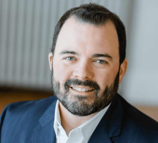 A professional portrait of a smiling man with a beard, wearing a suit and shirt, conveying confidence and approachability.