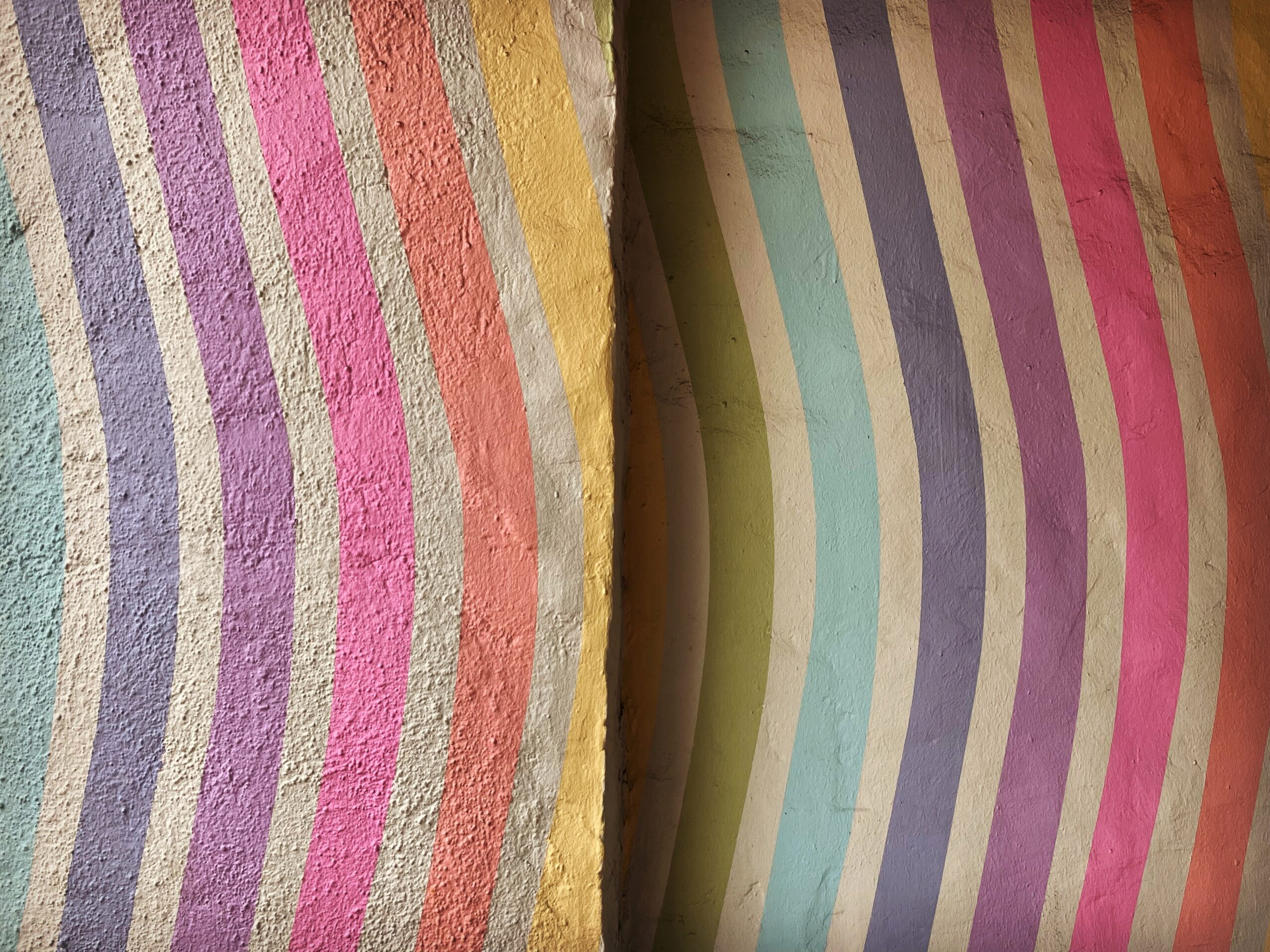 Curved vertical stripes in pastel colors, including purple, pink, yellow, blue, and green, are painted on a textured wall surface. A vertical crack or division runs through the middle of the image.