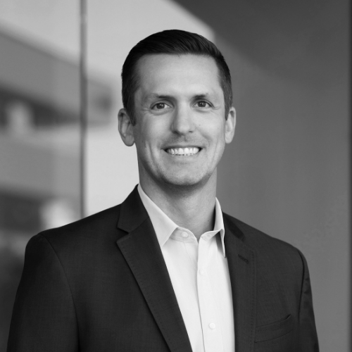 A man in a dark suit and light shirt smiles while standing indoors, with a blurred background featuring glass and architectural elements. The image is in black and white.