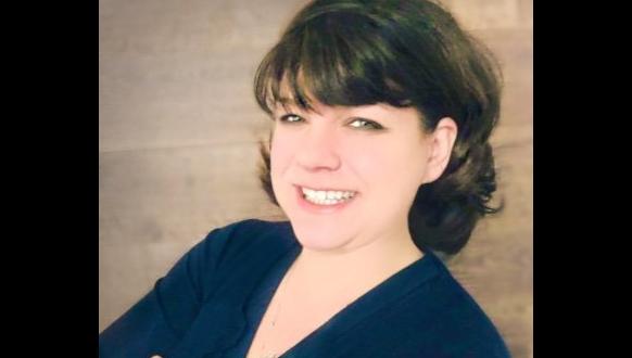 A woman with short, dark hair and bangs smiles at the camera. She is wearing a dark blue top and standing against a plain, light brown background.
