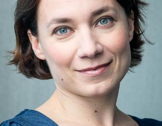 A woman with short brown hair and blue eyes is smiling softly. She is wearing a blue top and is positioned in front of a plain light-colored background.