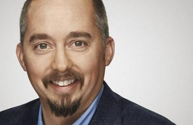 A smiling man with short brown hair, a trimmed beard and mustache, wearing a blue jacket and light blue shirt, posed against a plain light background.
