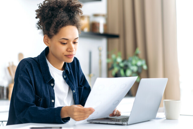 A woman sits at a table with a laptop, holding and looking at a sheet of paper. She appears focused, with a coffee mug and smartphone nearby in a bright, modern room.