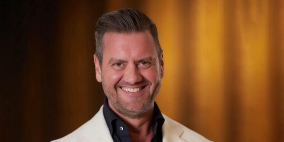 A man with short dark hair and a beard smiles, wearing a light-colored jacket over a dark shirt, posing in front of a blurred warm-toned background.